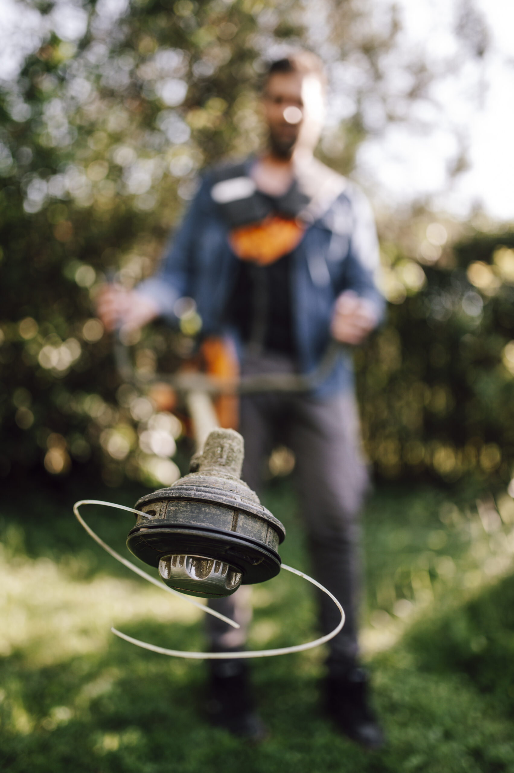 Close-up of a spinning string trimmer head with a person trimming grass in the background